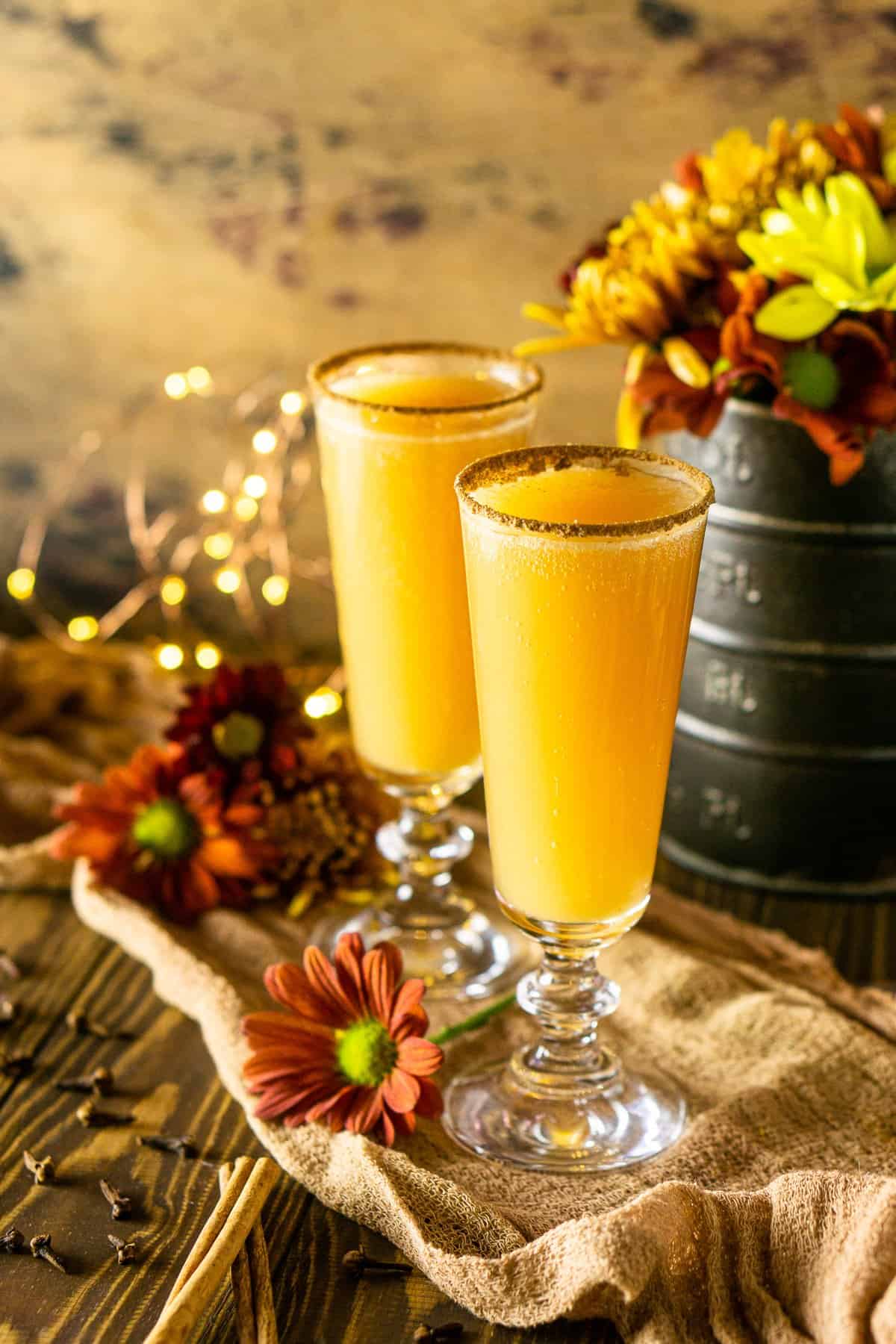 A side shot of two Thanksgiving mimosas on cheesecloth with cinnamon sticks and lights next to it.