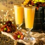 Two Thanksgiving mimosas with fall flowers and lights behind it with cinnamon sticks and cloves to the side.