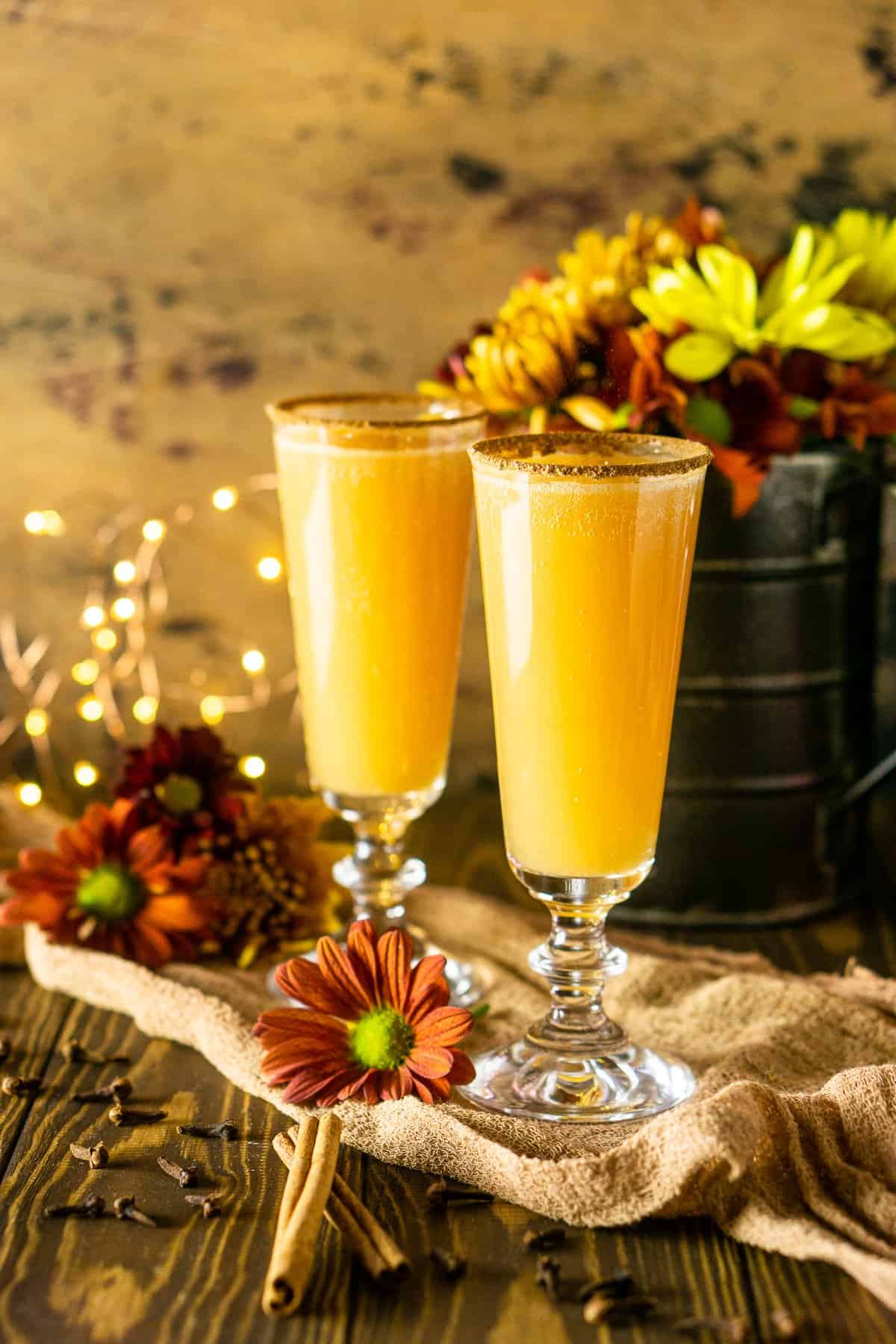 Two Thanksgiving mimosas with fall flowers and lights behind it with cinnamon sticks and cloves to the side.