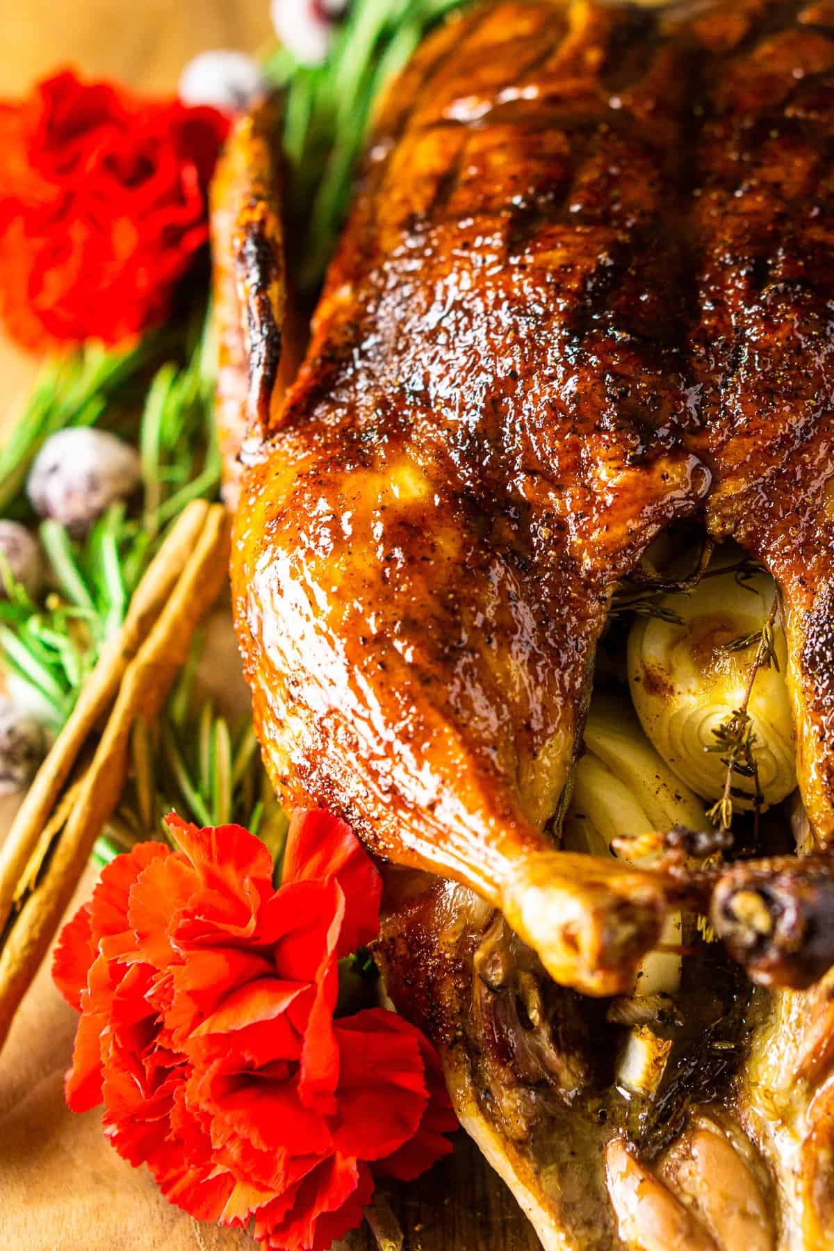 The finished Christmas roast duck on a wooden board surrounded by rosemary, cinnamon sticks, sugared cranberries and red flowers.