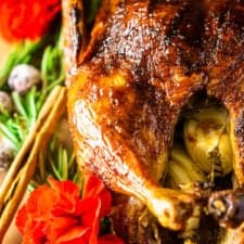 The finished Christmas roast duck on a wooden board surrounded by rosemary, cinnamon sticks, sugared cranberries and red flowers.