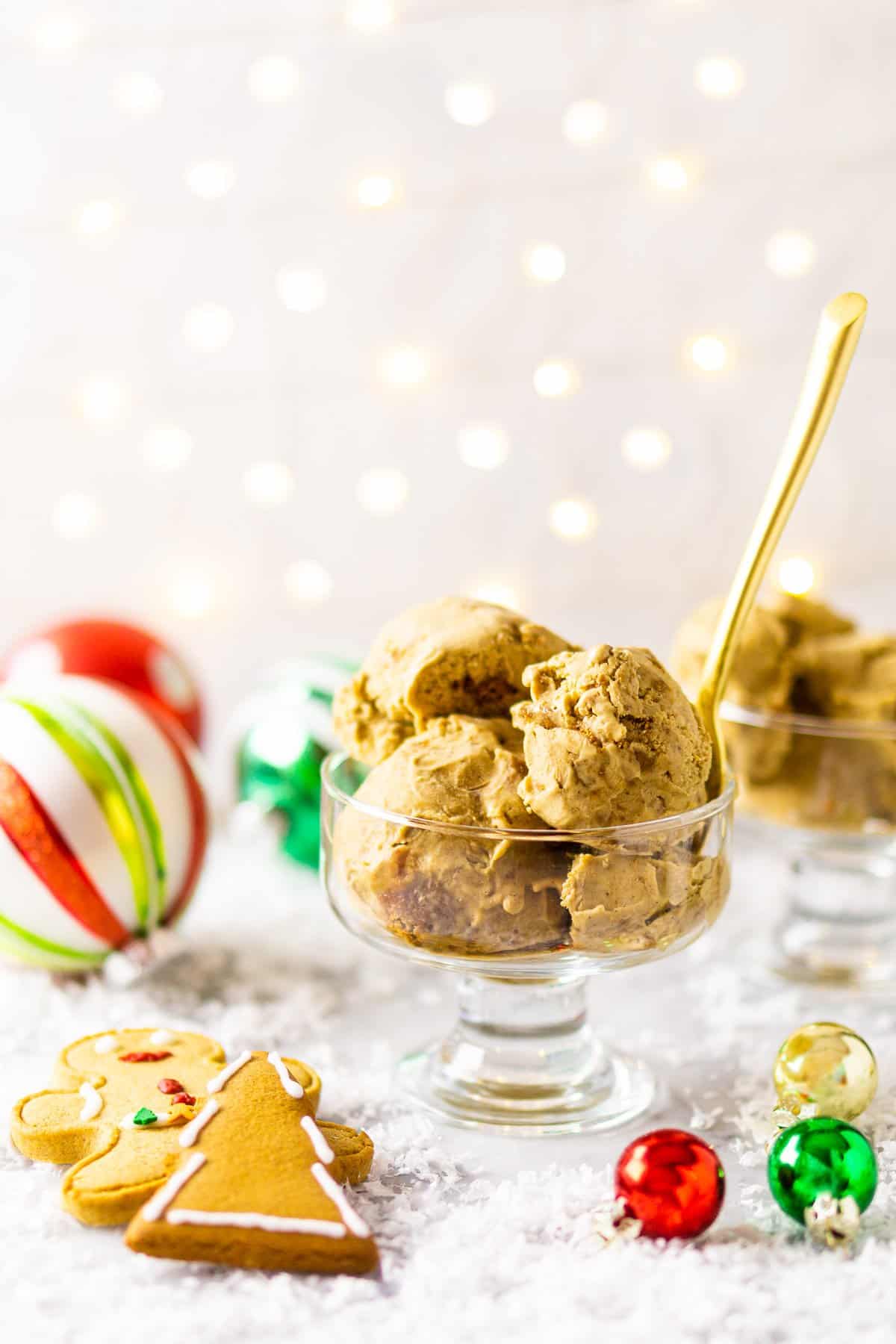 A glass bowl of gingerbread ice cream with ornaments and gingerbread cookies around it with a gold spoon sticking out of the bowl.