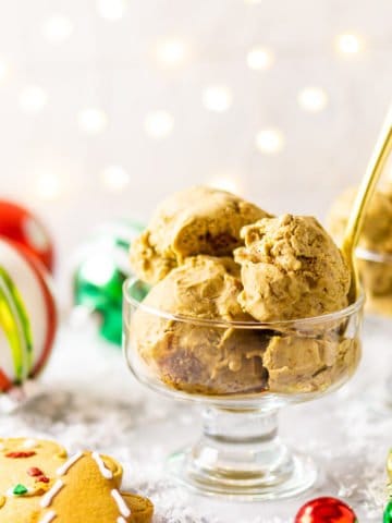 A glass bowl of gingerbread ice cream with ornaments and gingerbread cookies around it with a gold spoon sticking out of the bowl.