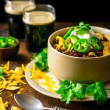 A bowl of authentic Texas chili with cilantro, cheese and two stouts in the background.