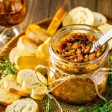 A wooden plate with a mason jar filled with bacon jam surrounded by bread slices, fresh thyme and a glass of bourbon.