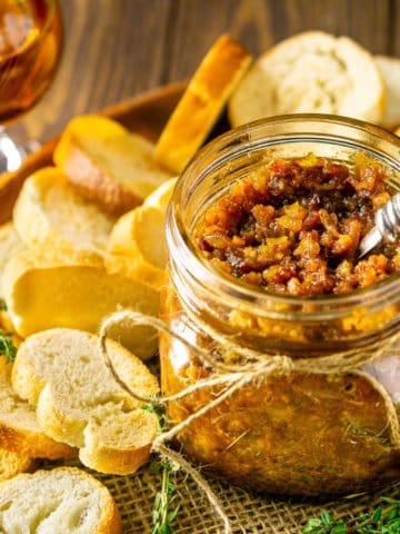 A wooden plate with a mason jar filled with bacon jam surrounded by bread slices, fresh thyme and a glass of bourbon.