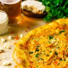 The Buffalo chicken pizza on parchment paper with ranch and a beer to the side with a slab of blue cheese and fresh parsley.