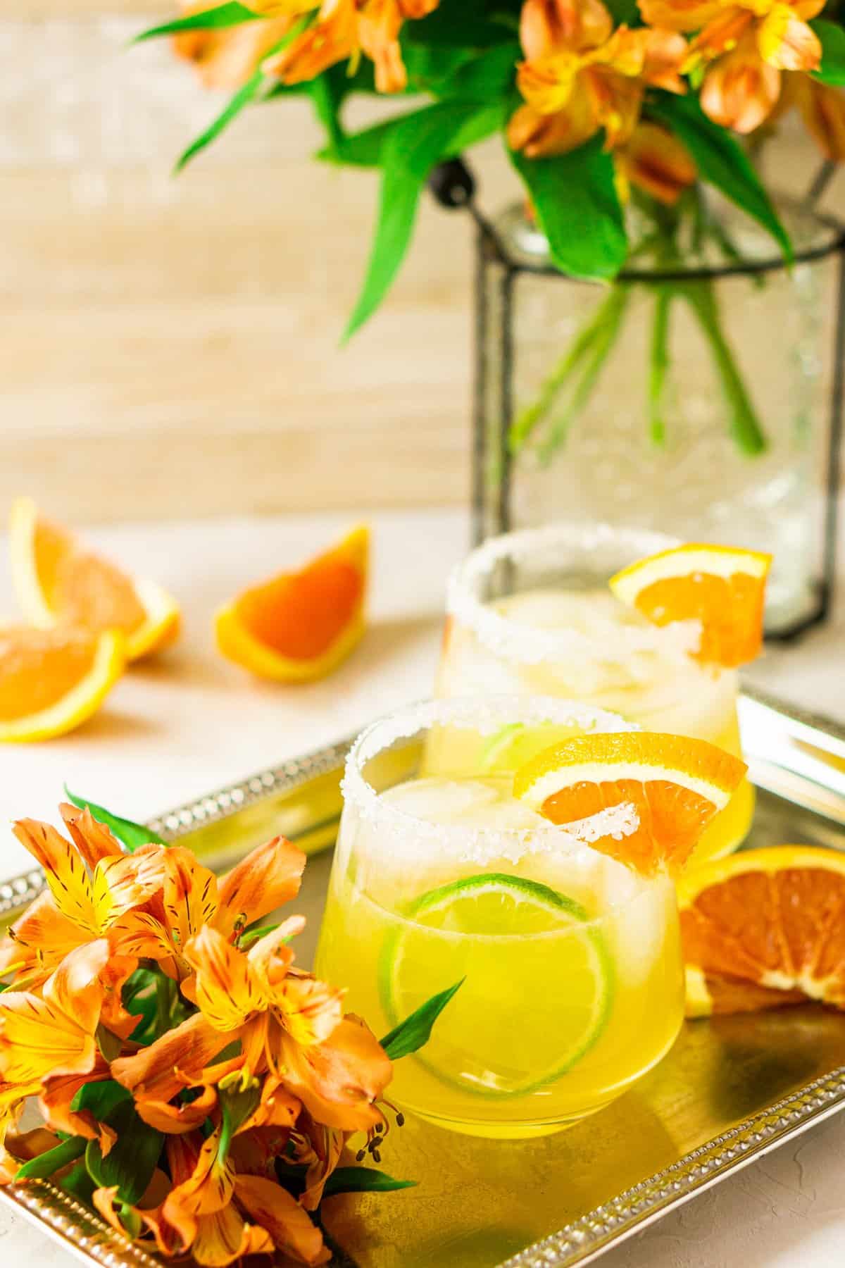 Two orange margaritas on a silver platter with flowers on the side.