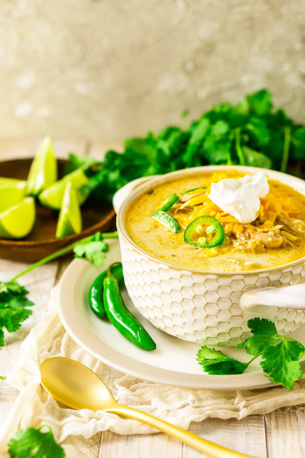 A bowl of the creamy white chicken chili with lime slices and a bunch of cilantro in the background with a gold spoon to the side.