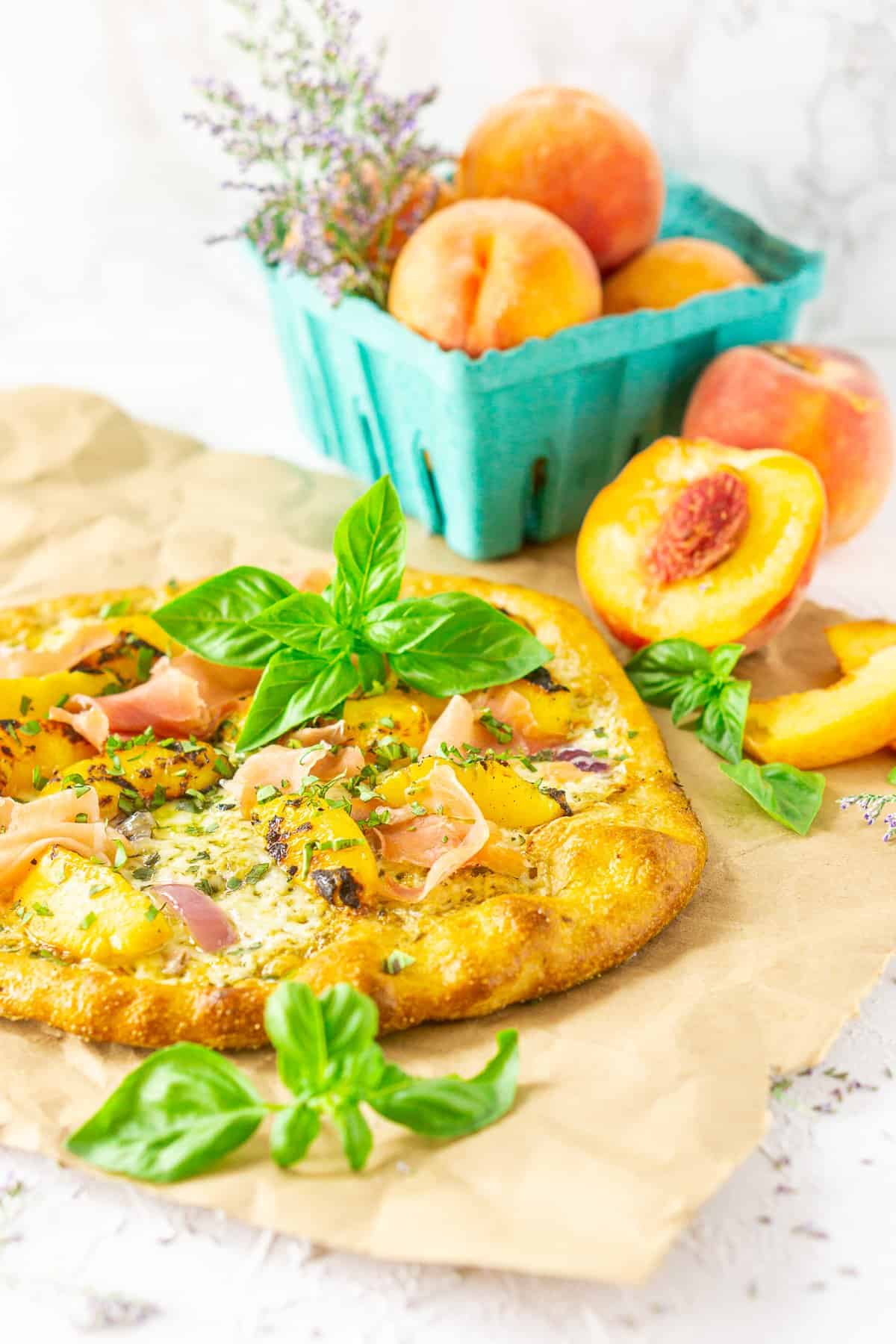 A grilled peach pizza on parchment paper with fresh peaches and purple flowers.