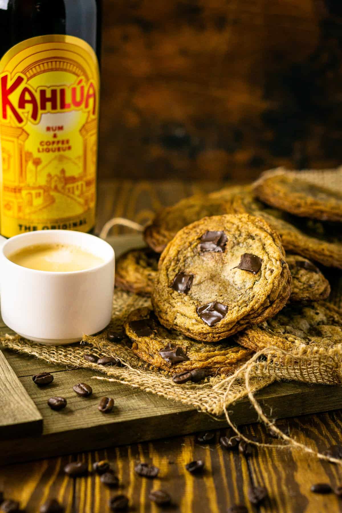 A pile of espresso and Kahlua cookies on a wooden tray with burlap and a cup of espresso and bottle of Kahlua next to it.