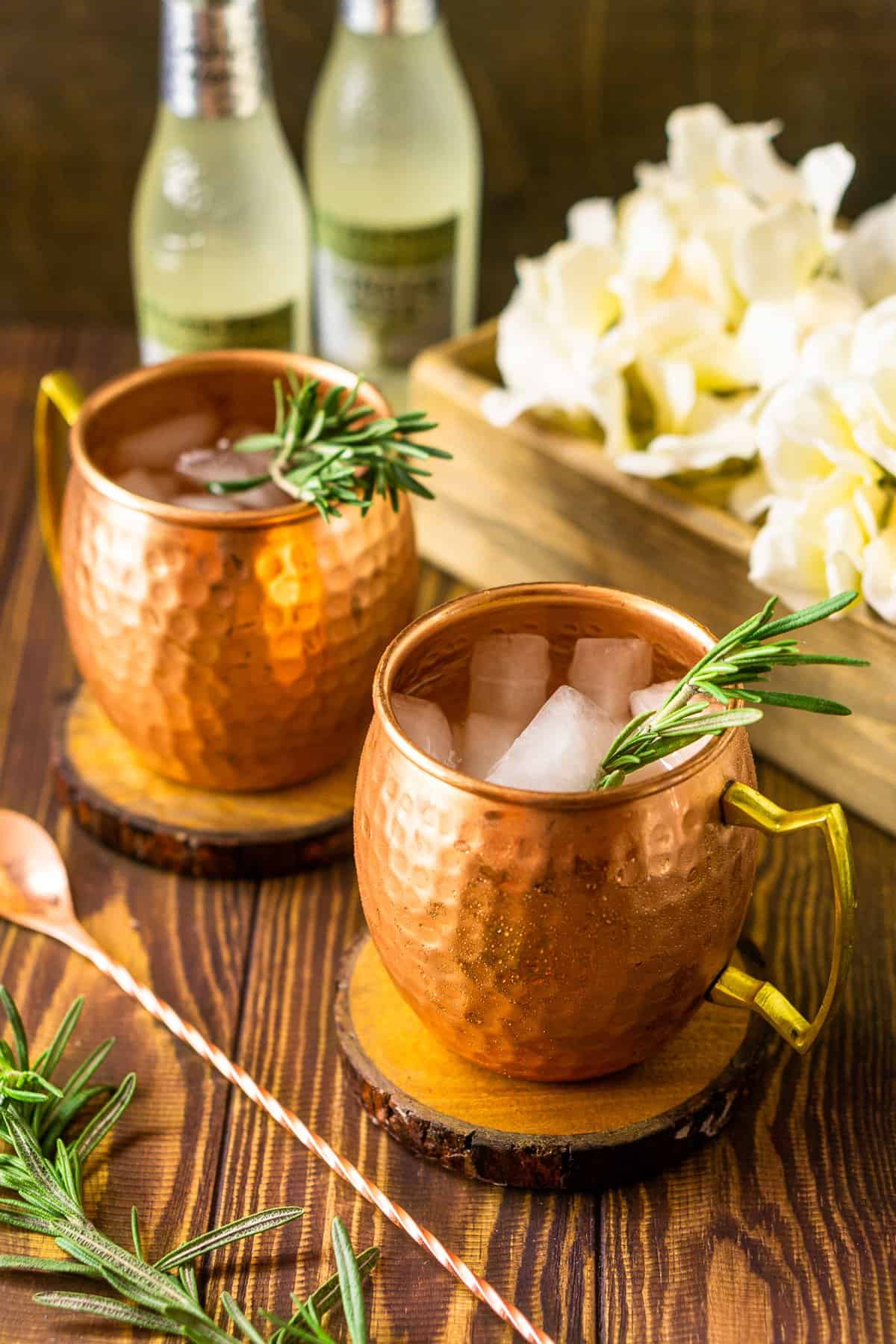 Two rosemary Kentucky mules with a copper bar spoon to the left and a box of white flowers in the background.