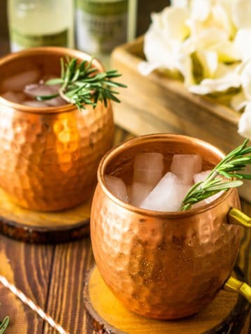 Two rosemary Kentucky mules with a copper bar spoon to the left and a box of white flowers in the background.