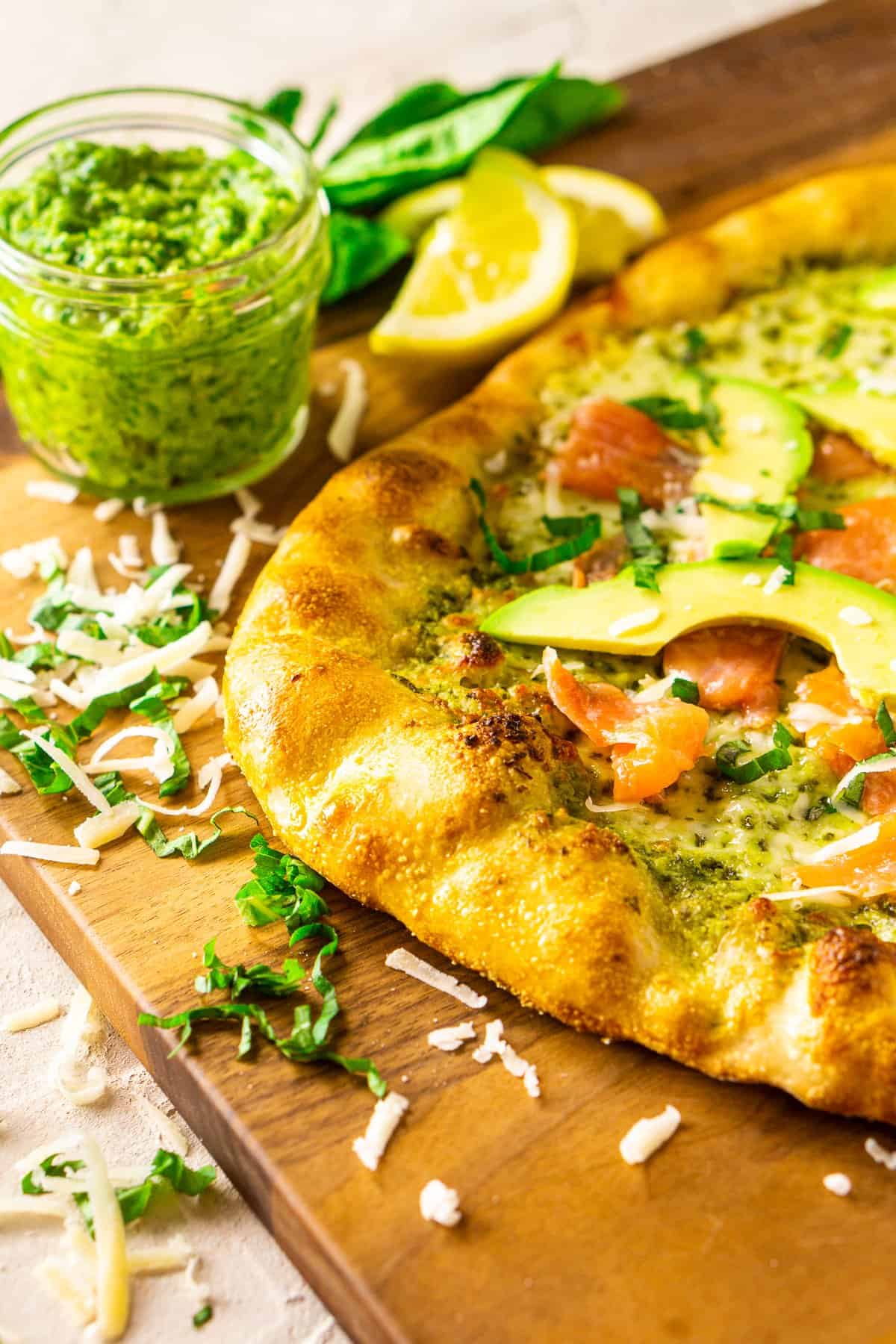 The smoked salmon and avocado pizza with pesto on a wooden cutting board with a jar of pesto, herbs and cheese around it.