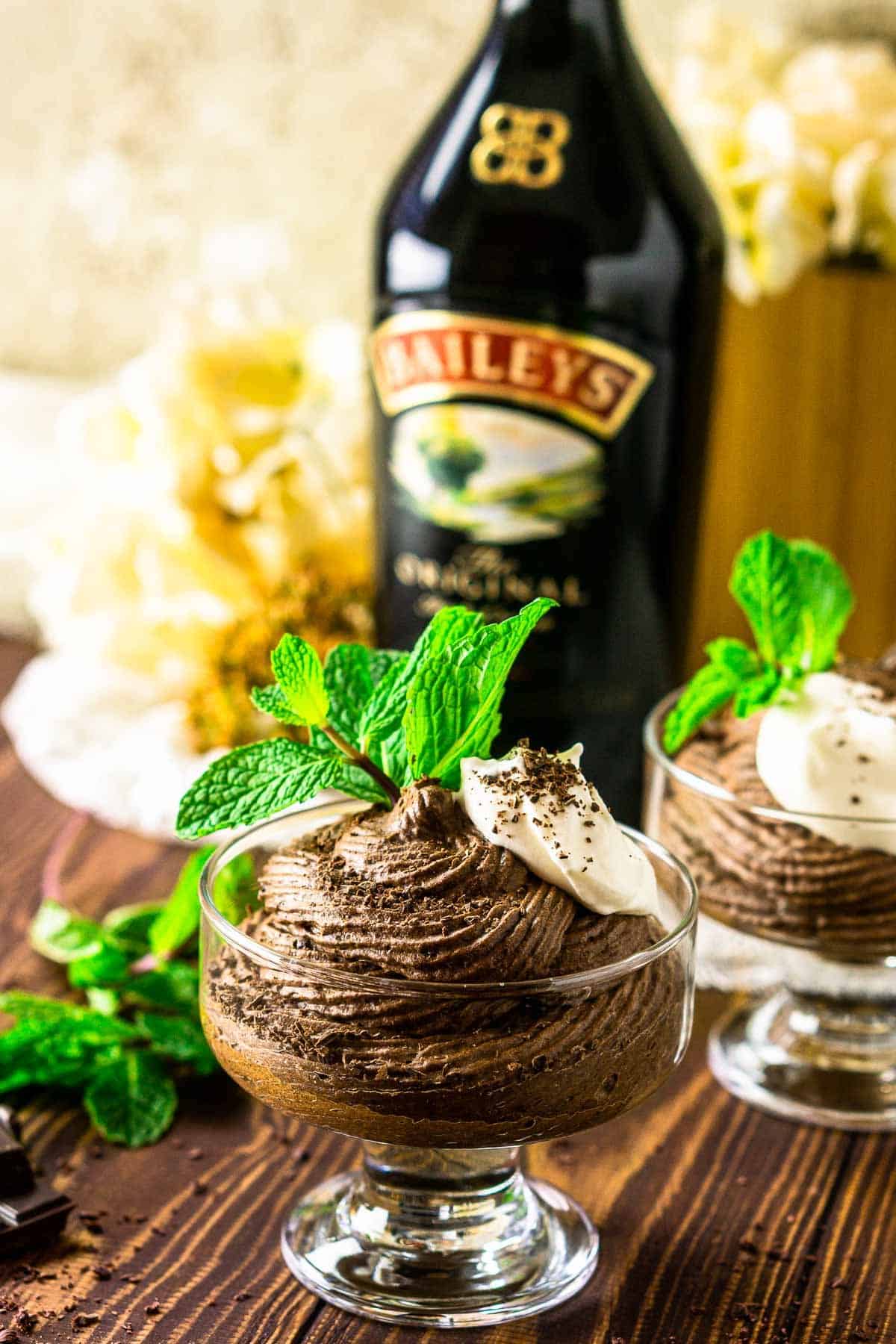 A cup of Baileys mousse with a bottle of Irish cream behind it with a sprig of mint to the side.