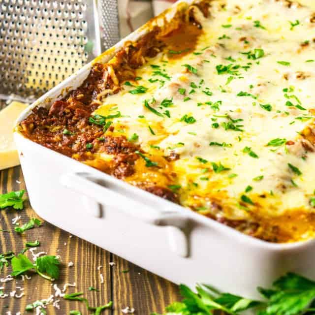 A pan of meatball lasagna with fresh parsley and a cheese grate with a slab of Parmesan.