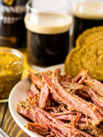 A plate of shredded slow cooker Guinness corned beef with mustard on the side and two beers in the background.