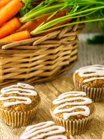 The carrot cake muffins in front of a basket with fresh carrots in it.