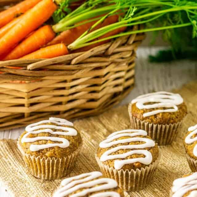 The carrot cake muffins in front of a basket with fresh carrots in it.