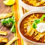 Two bowls of Instant Pot chicken enchilada soup on a colorful napkin with cilantro, tortilla chip strips and lime slices to the side.