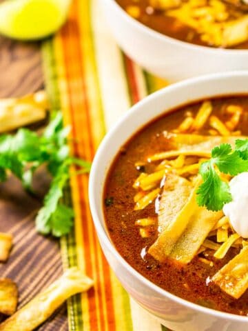 Two bowls of Instant Pot chicken enchilada soup on a colorful napkin with cilantro, tortilla chip strips and lime slices to the side.