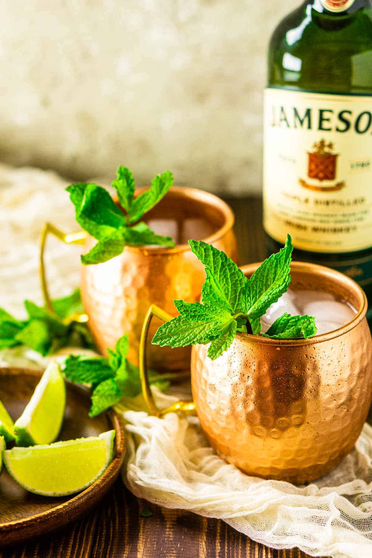 Two Irish mule cocktails on a cream-colored clothe with limes to the side and a bottle of Jameson behind them.