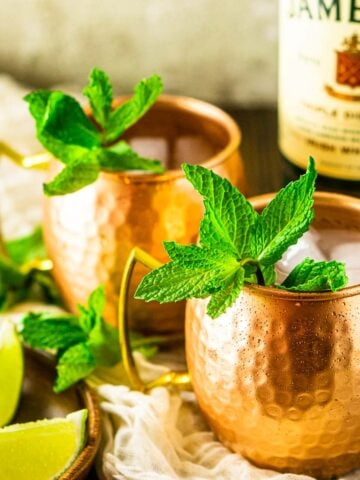 Two Irish mule cocktails on a cream-colored clothe with limes to the side and a bottle of Jameson behind them.