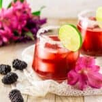 Two fresh blackberry margaritas on lace with pink and purple flowers in the background.
