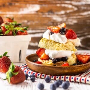 A blueberry-strawberry shortcake on a wooden plate and patriotic clothe with berries surrounding it.