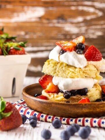 A blueberry-strawberry shortcake on a wooden plate and patriotic clothe with berries surrounding it.