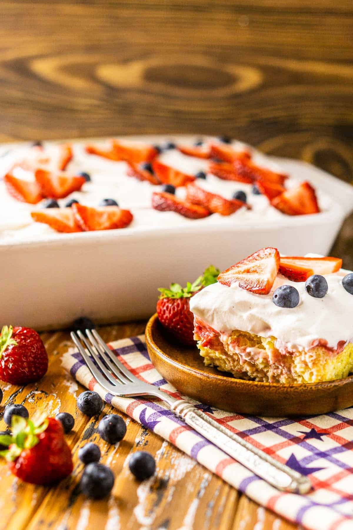 A slice of the 4th of July poke cake on a wooden plate with berries to the side.