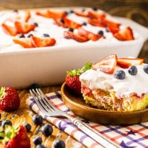 A slice of the 4th of July poke cake on a wooden plate with berries to the side.
