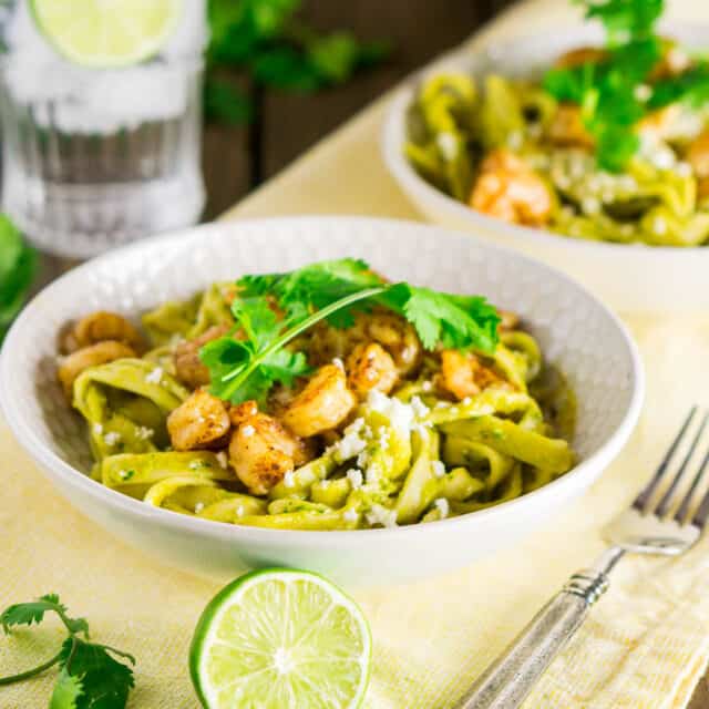 Two bowls of spicy avocado pasta with tequila shrimp on a yellow napkin with cilantro to the side.