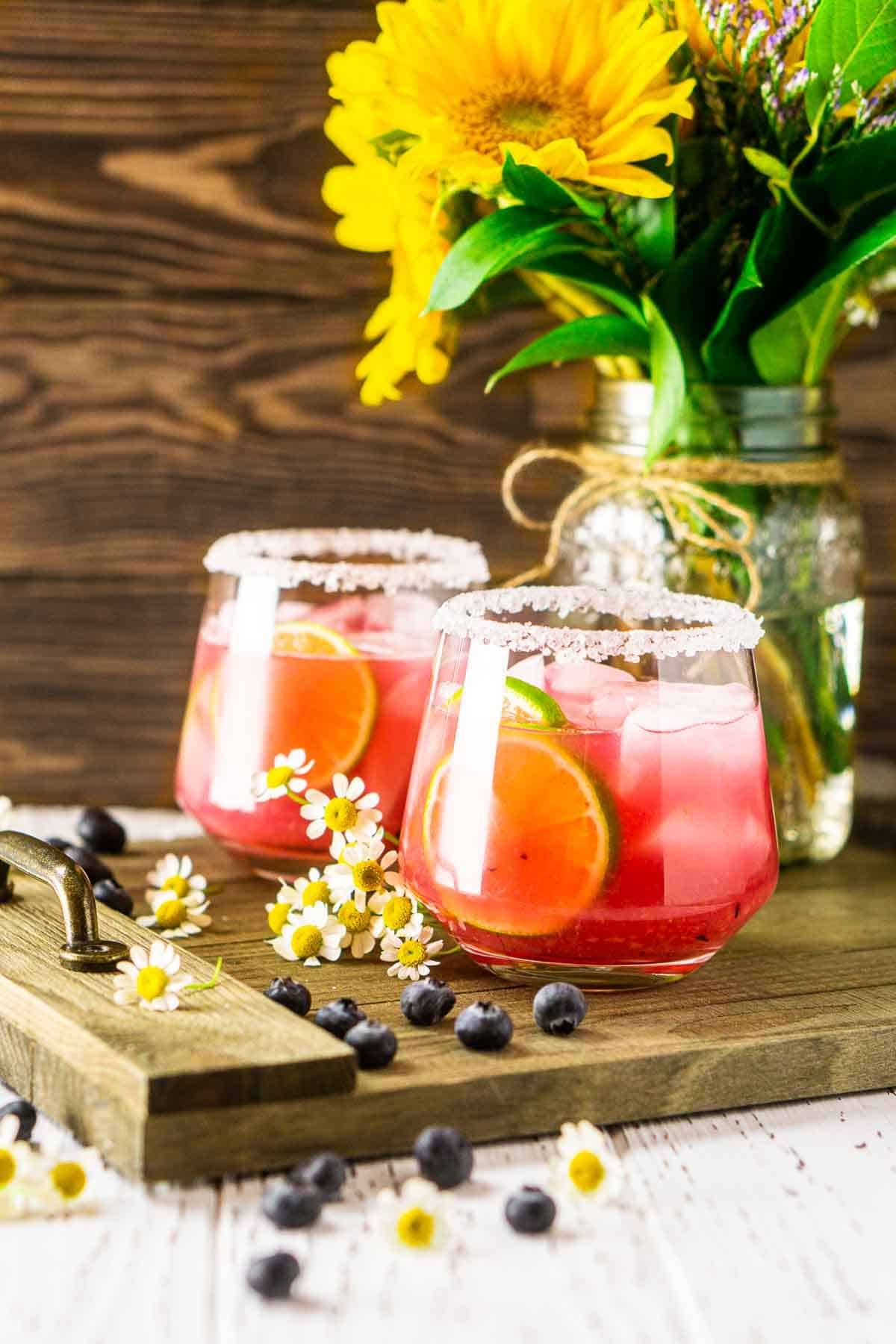 Two fresh blueberry margaritas with flowers and blueberries to the side on a wooden tray.