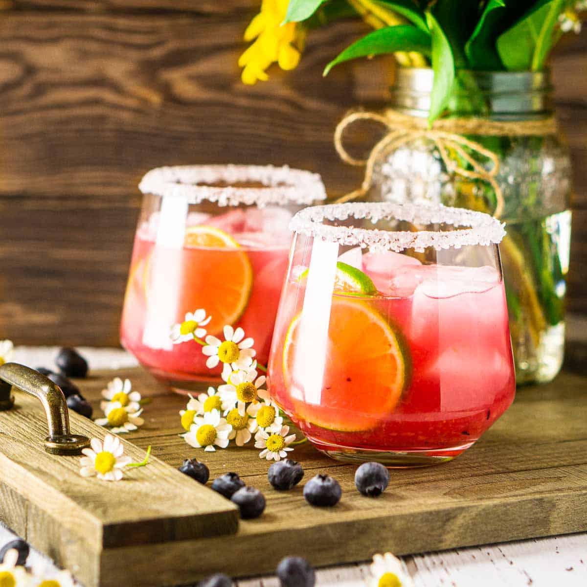 Two blueberry margaritas on a wooden tray with fresh blueberries and flowers surrounding them.