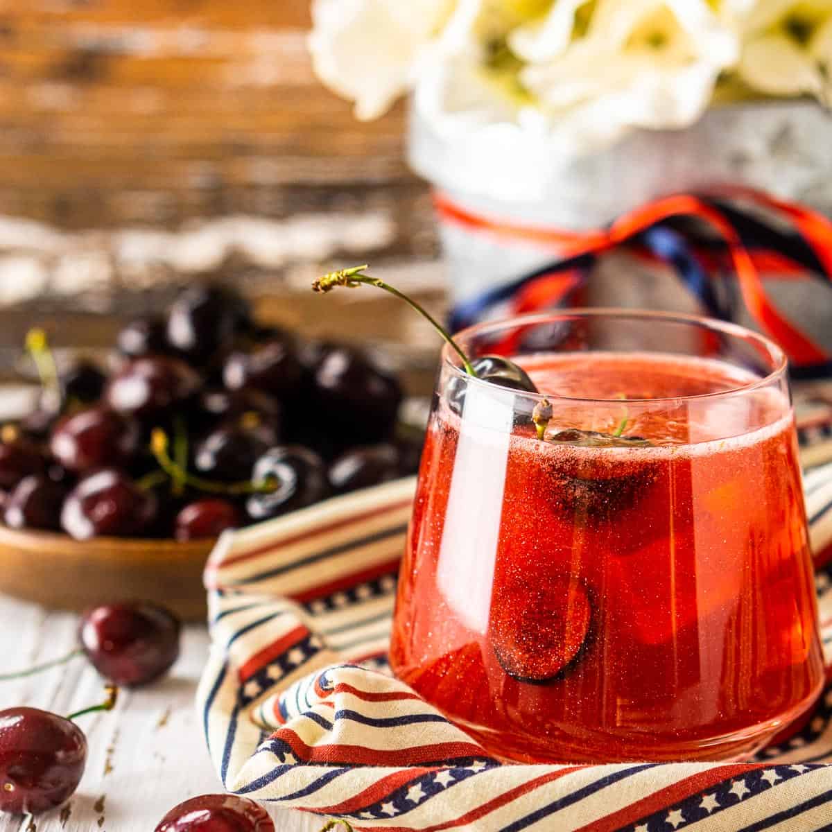 A Cherry Firecracker cocktail on patriotic clothe with cherries around it.