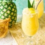 Two painkiller cocktails on a straw placemat with a pineapple and coconut to the side.