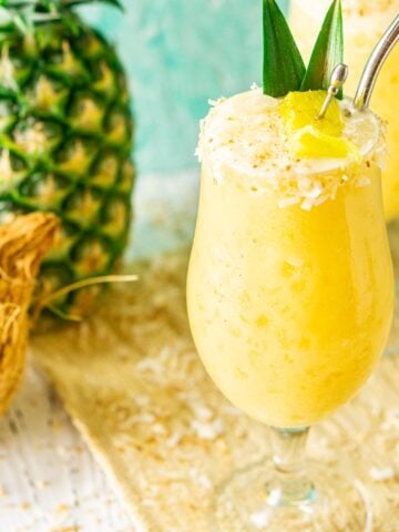 Two painkiller cocktails on a straw placemat with a pineapple and coconut to the side.