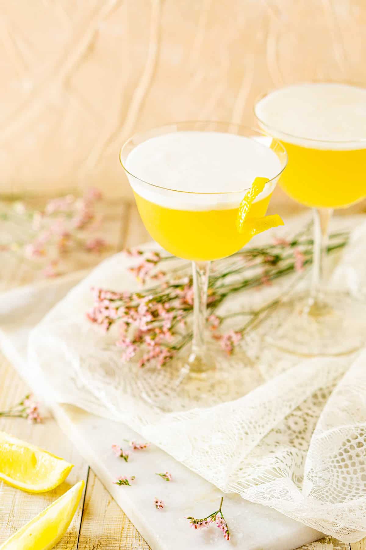 Two tequila sour cocktails on white lace with flowers and lemon slices around them.