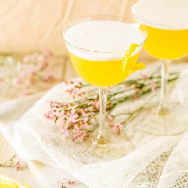 Two tequila sour cocktails on white lace with flowers and lemon slices around them.