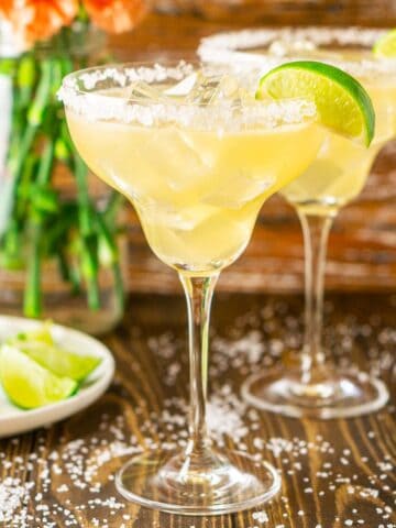 Two Cadillac margaritas with salt around them and limes and flowers to the side.