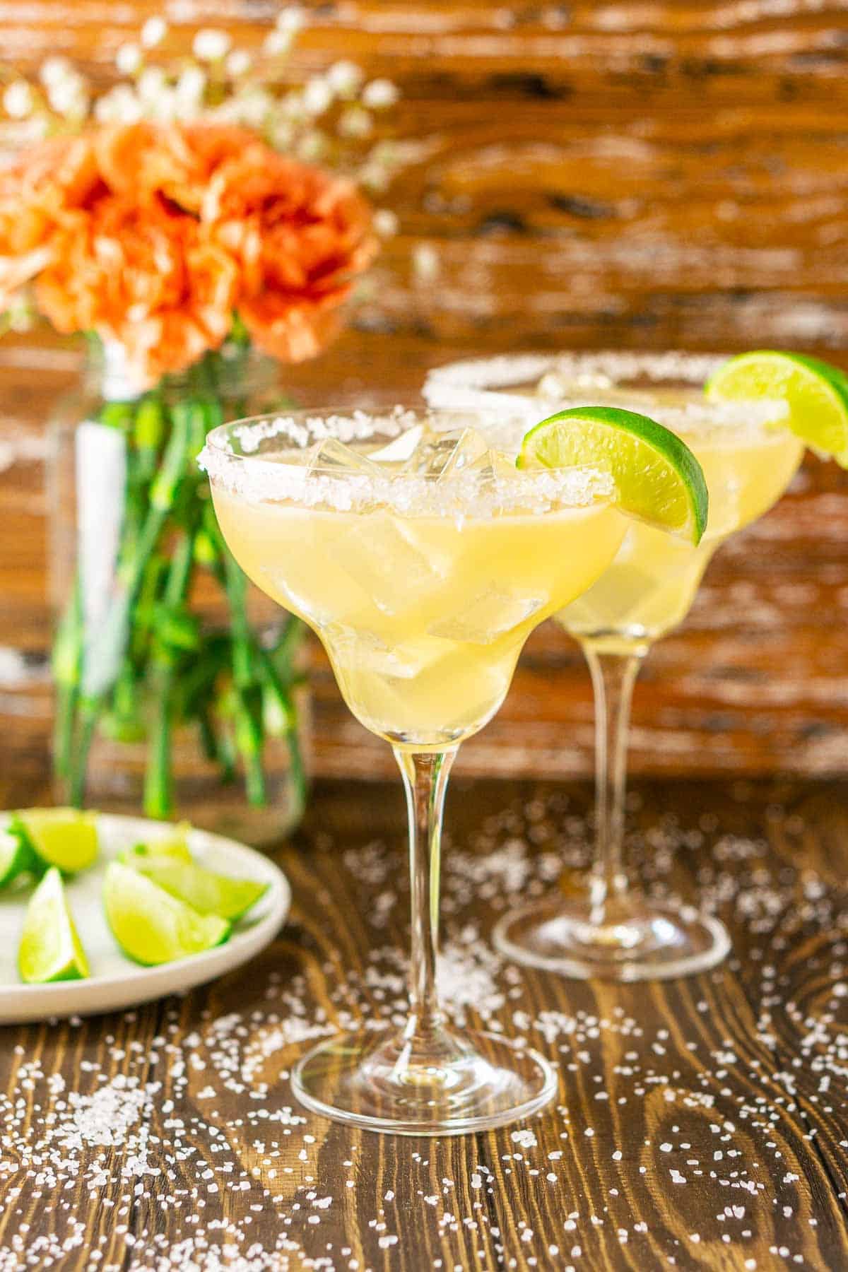 Two Cadillac margaritas with salt around them and limes and flowers to the side.