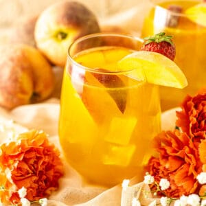 Two glasses of peach sangria with flowers in front of them and fresh peaches in the background.
