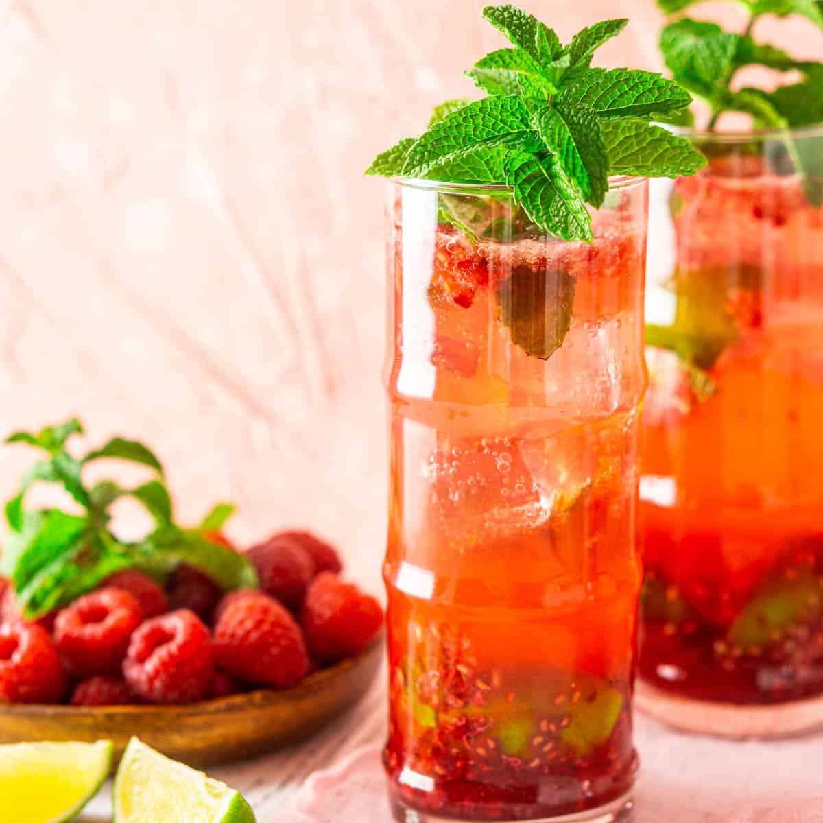 A raspberry mojito with fresh berries and lime wedges surrounding it against a pink background.