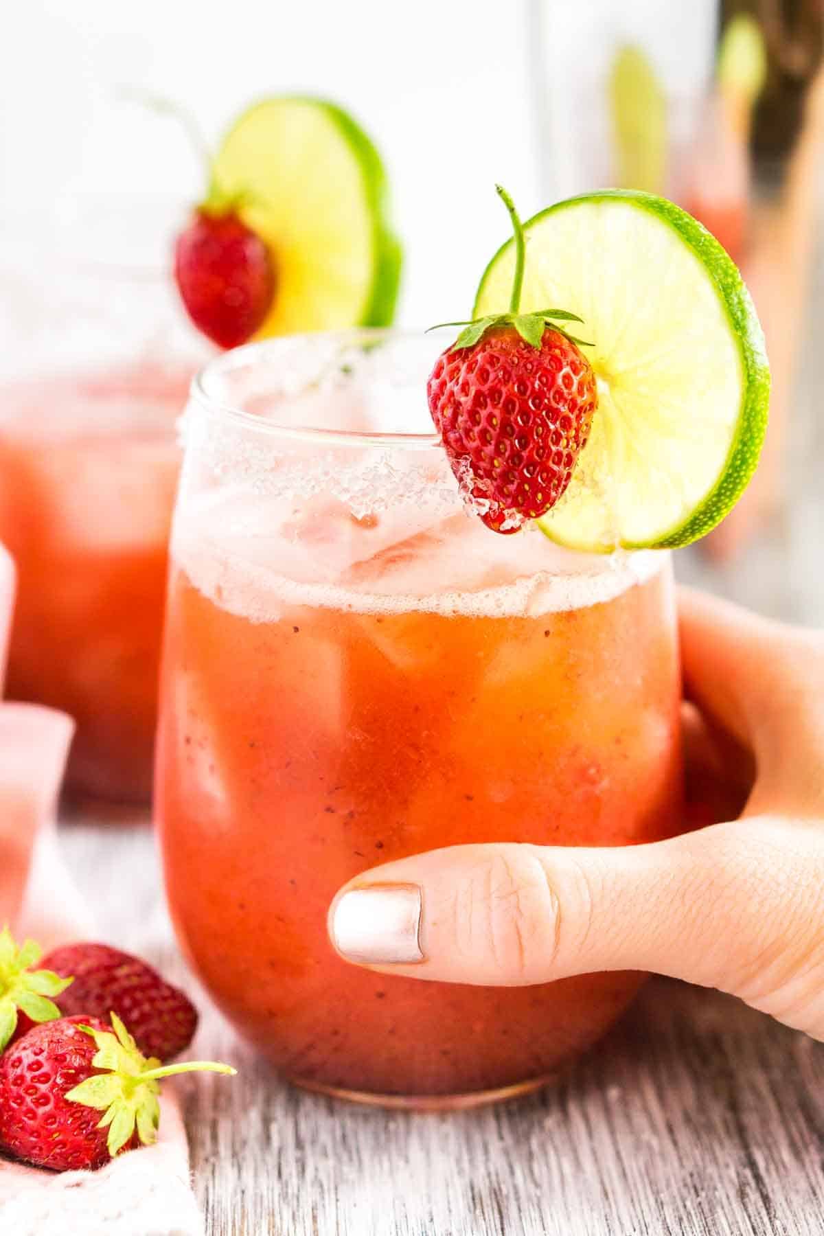 A hand holding the roasted strawberry margarita with fresh strawberries to the left.