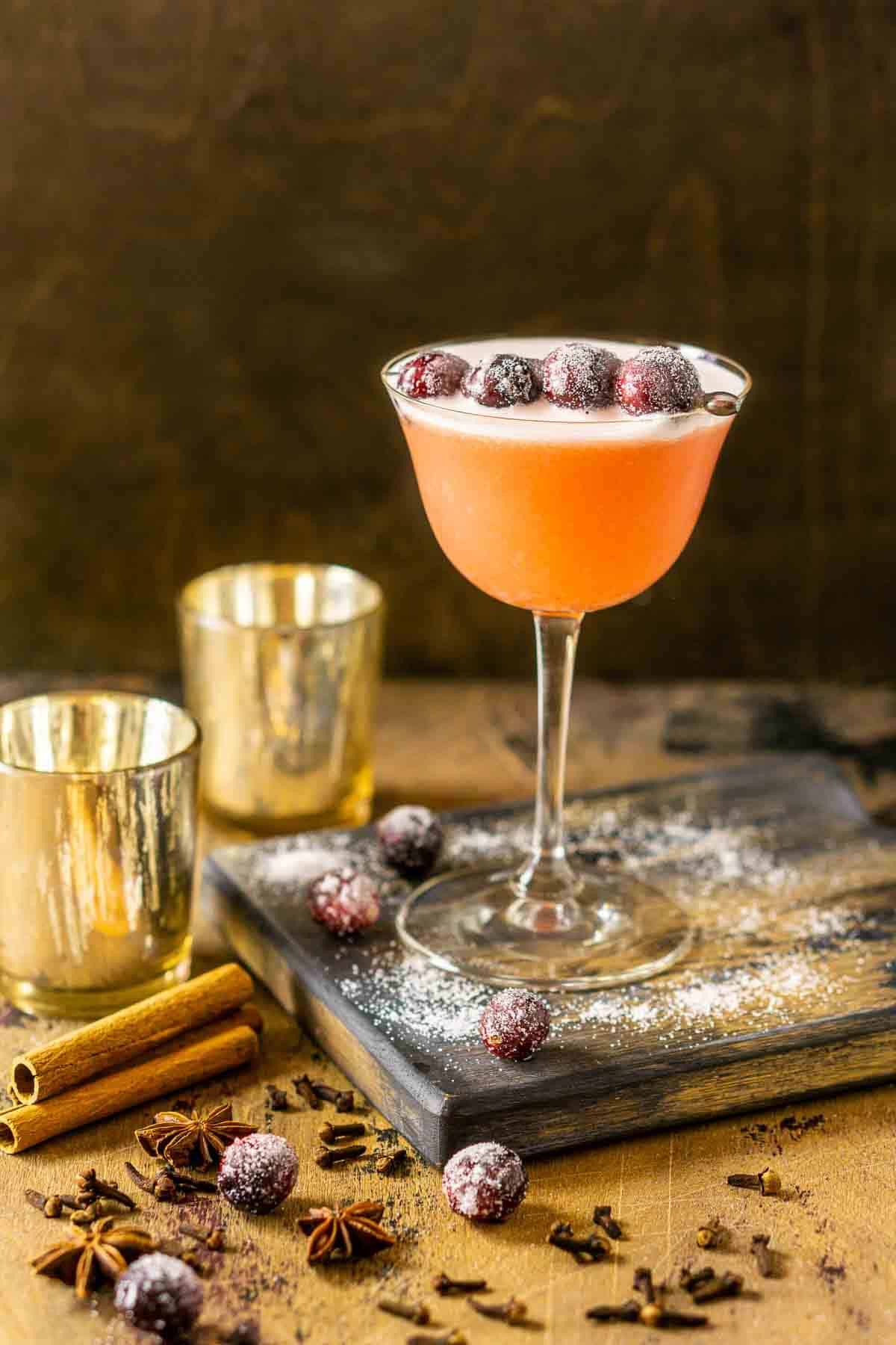A spiced cranberry bourbon sour on a wooden board with candles and sugared cranberries around it.