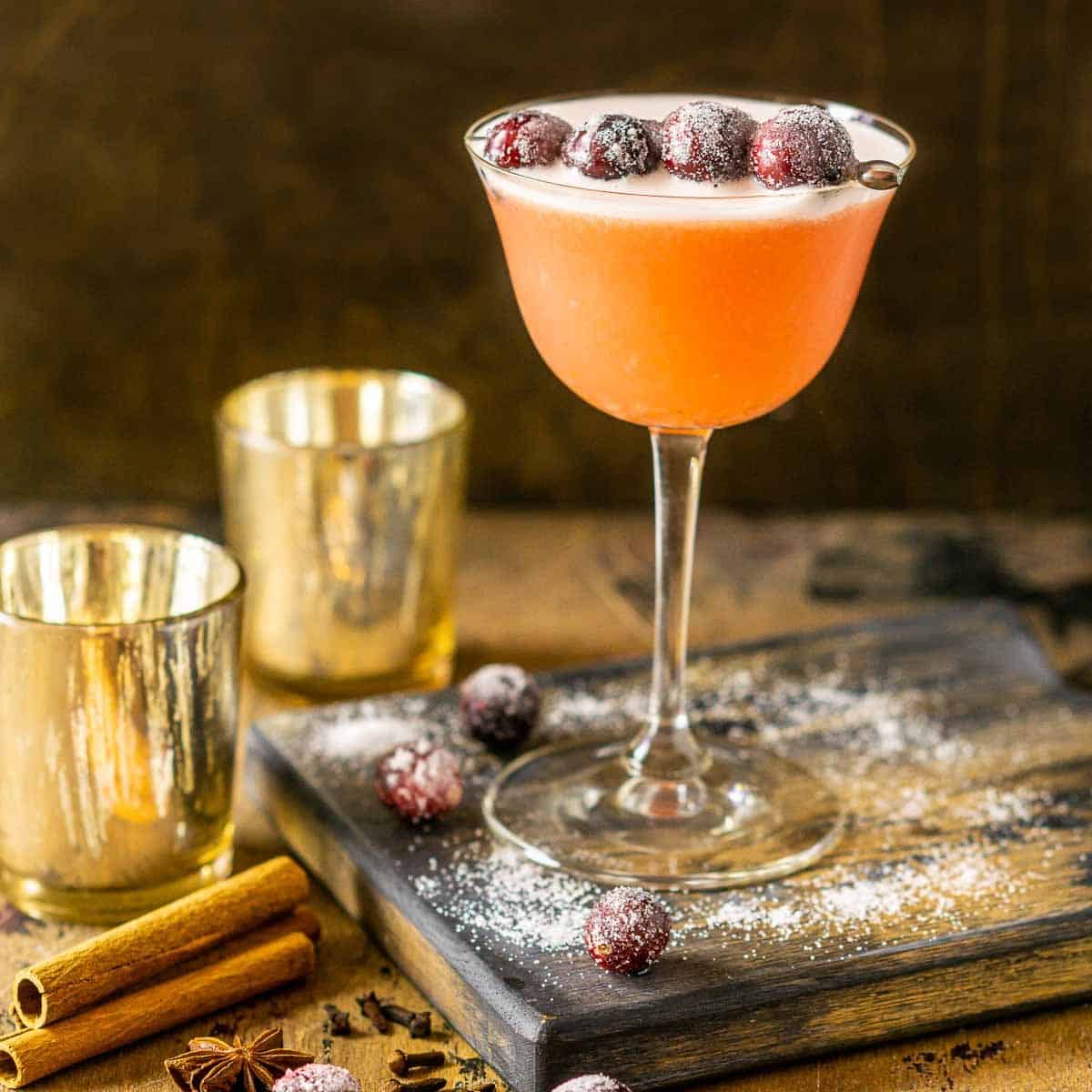 A cranberry bourbon sour with candles, sugared cranberries and spices to the side.