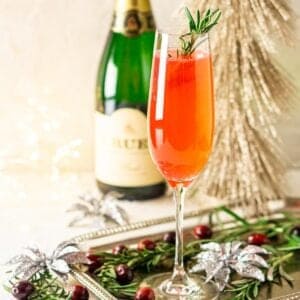 A cranberry French 75 on a silver platter with rosemary and cranberries around it with a bottle of Champagne in the background.