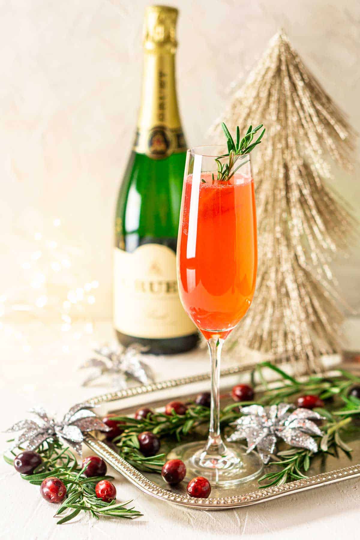 A cranberry French 75 on a silver platter with rosemary and cranberries around it with a bottle of Champagne in the background.
