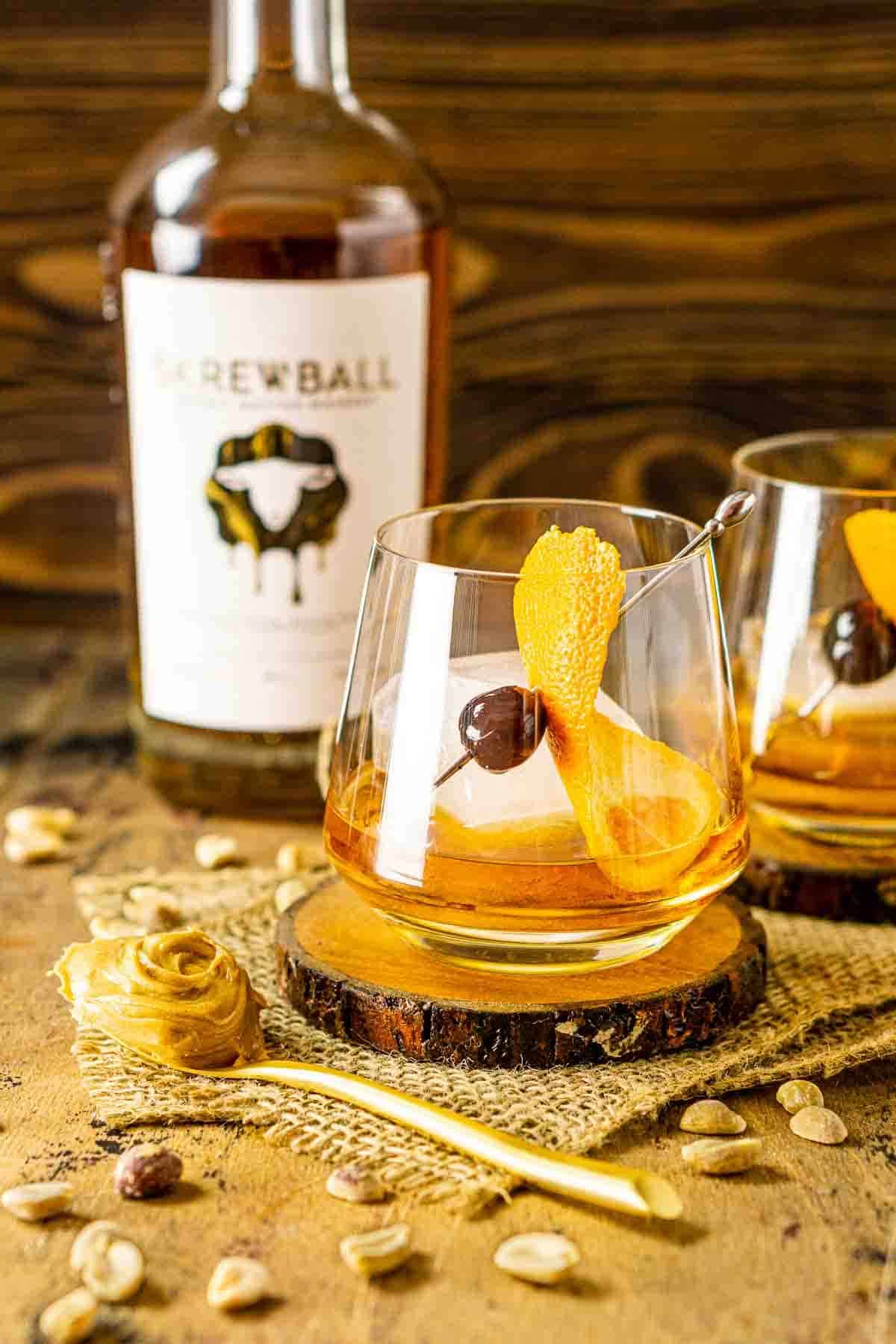 Two peanut butter old fashioned cocktails on a wooden coaster with a bottle of whiskey in the background.